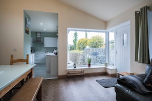 A modern kitchen with white cabinets and a stainless steel range hood, a dining area with a wooden bench and table, and a cozy seating area with a leather sofa and a window with a view of a stone wall and greenery. - Five Acres - Sleeps 8 - Private Hot Tub - Pet Friendly - Conwy (Conwy)