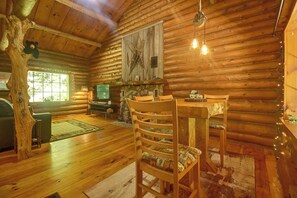 Het interieur van een blokhut met een eettafel, stoelen, een open haard en een groot raam.