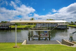 A covered boat lift structure with a grassy area in the foreground and a body of water with boats in the background. - Canal-Front Home w/ Boat Lift & Pool in Cape Coral (Cape Coral)