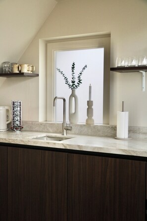 A kitchen with a sink, a faucet, a vase with a plant, and a shelf with cups and glasses.