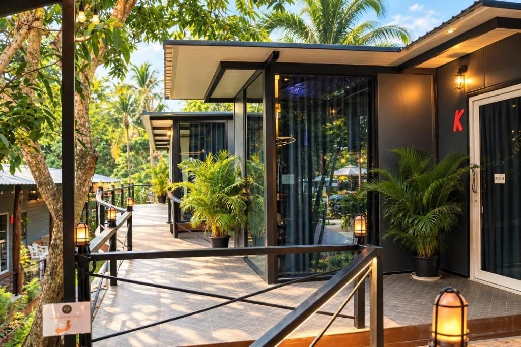 Exterior moderno de un hotel con entrada de vidrio, mobiliario exterior y plantas en macetas.