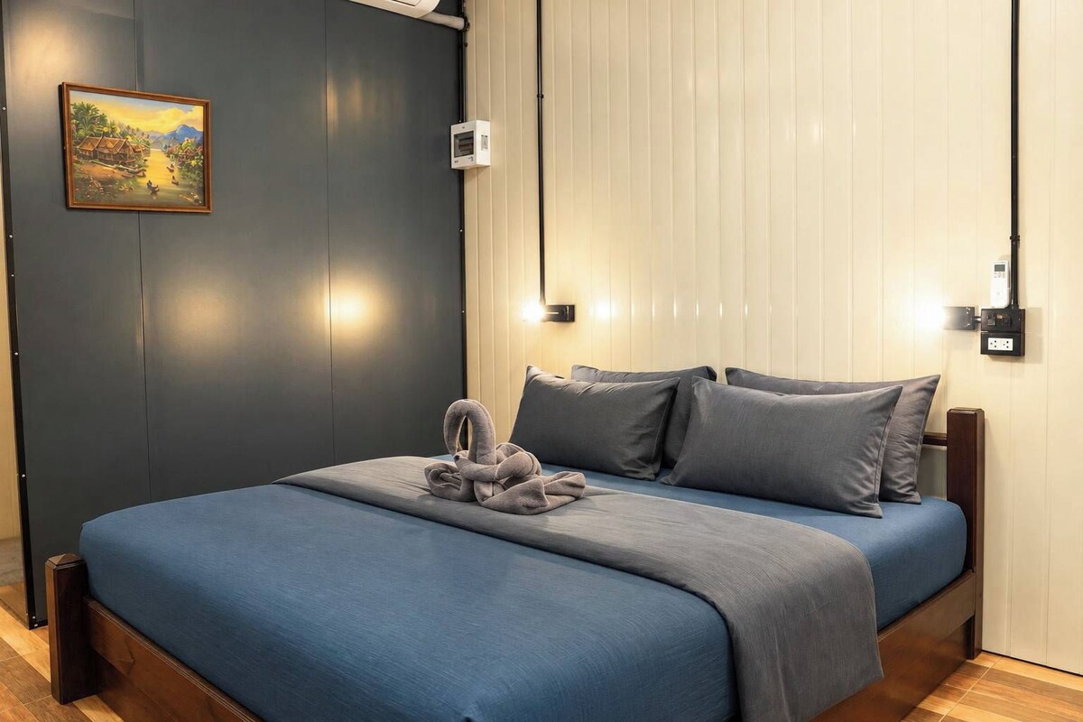 A bedroom with a bed, pillows, a wall-mounted light, a power outlet, and a painting.