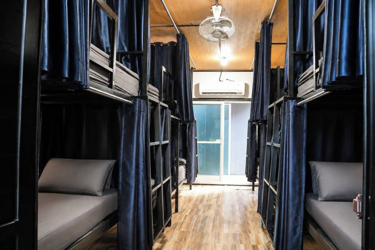 A train carriage interior with bunk beds, curtains, a ceiling fan, and an air conditioning unit.