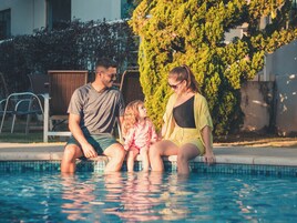 Una familia de tres junto a una piscina, con un seto y sillas al fondo.