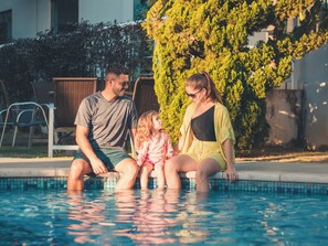 Una familia de tres junto a una piscina, con un seto y sillas al fondo.