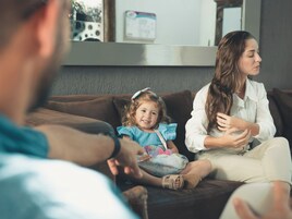 Una sala de estar con un sofá marrón, una niña con un vestido azul y un adulto con una camisa blanca.