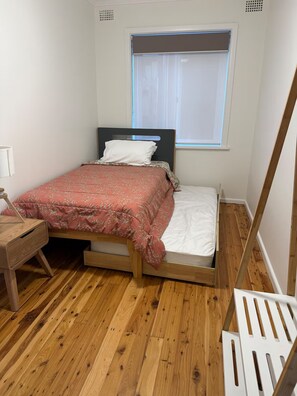 A small, single-bed room with a wooden floor, a window with blinds, a bedside table, and a chair. - Cosy 3-Bedroom Coastal Retreat with Ocean Views Opposite a Patrolled Beach (Bulli)