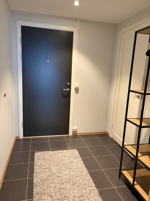 Et værelse med en mørk dør, en hvid væg, et flisegulv og en reol i træ.