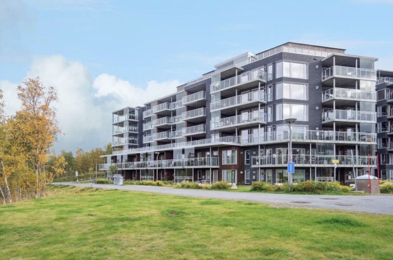 A multi-story residential building with balconies, a flat roof, and a grassy area in front.