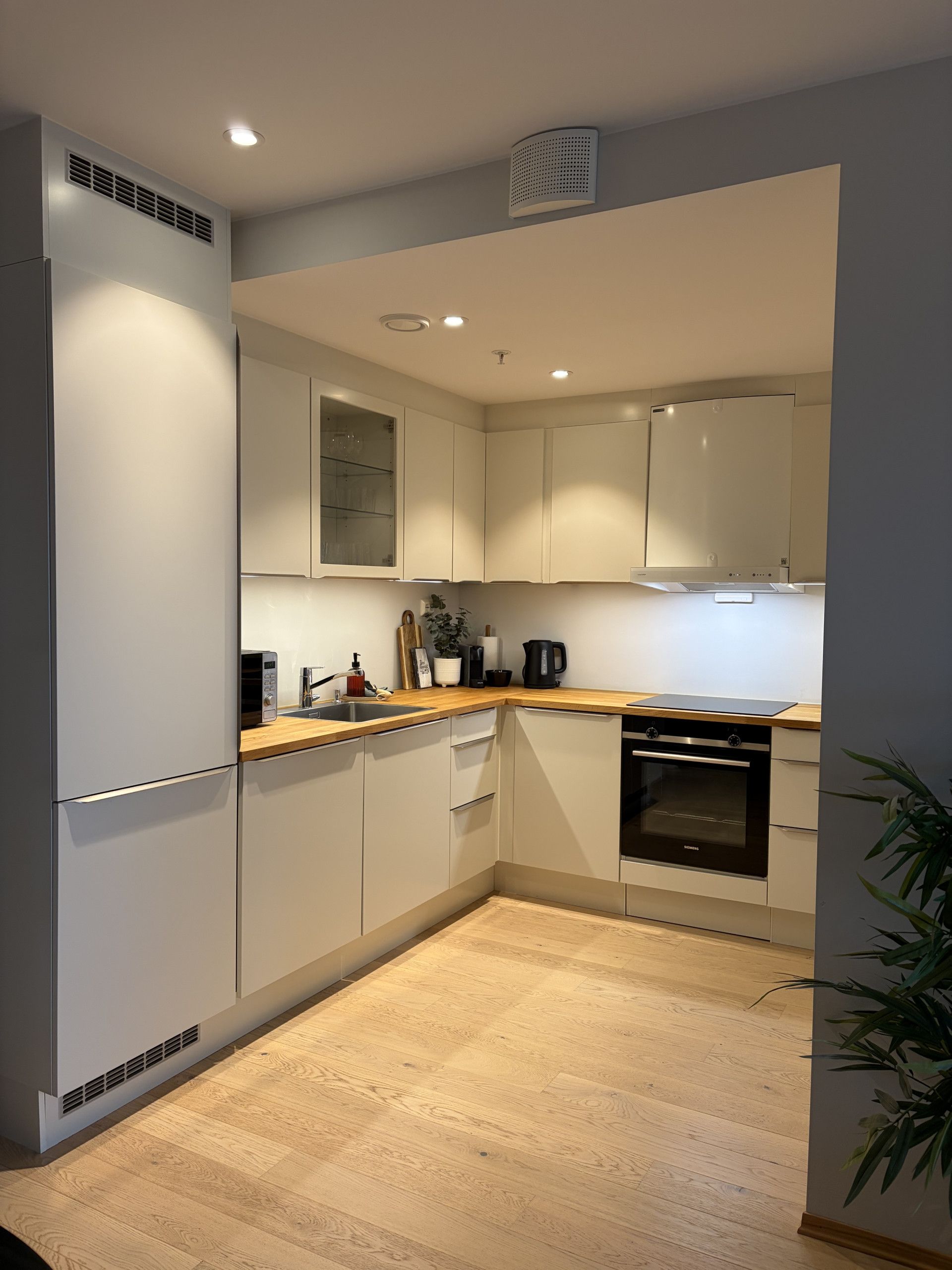 Een moderne keuken met witte kasten, een houten aanrechtblad, een inbouwoven en een ventilatierooster aan de muur.