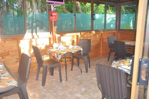 An outdoor patio area with wooden walls, a covered seating area with wicker chairs and tables, and a view of greenery through the windows. - Hostal y Cabanas Anakena (Hanga Roa)