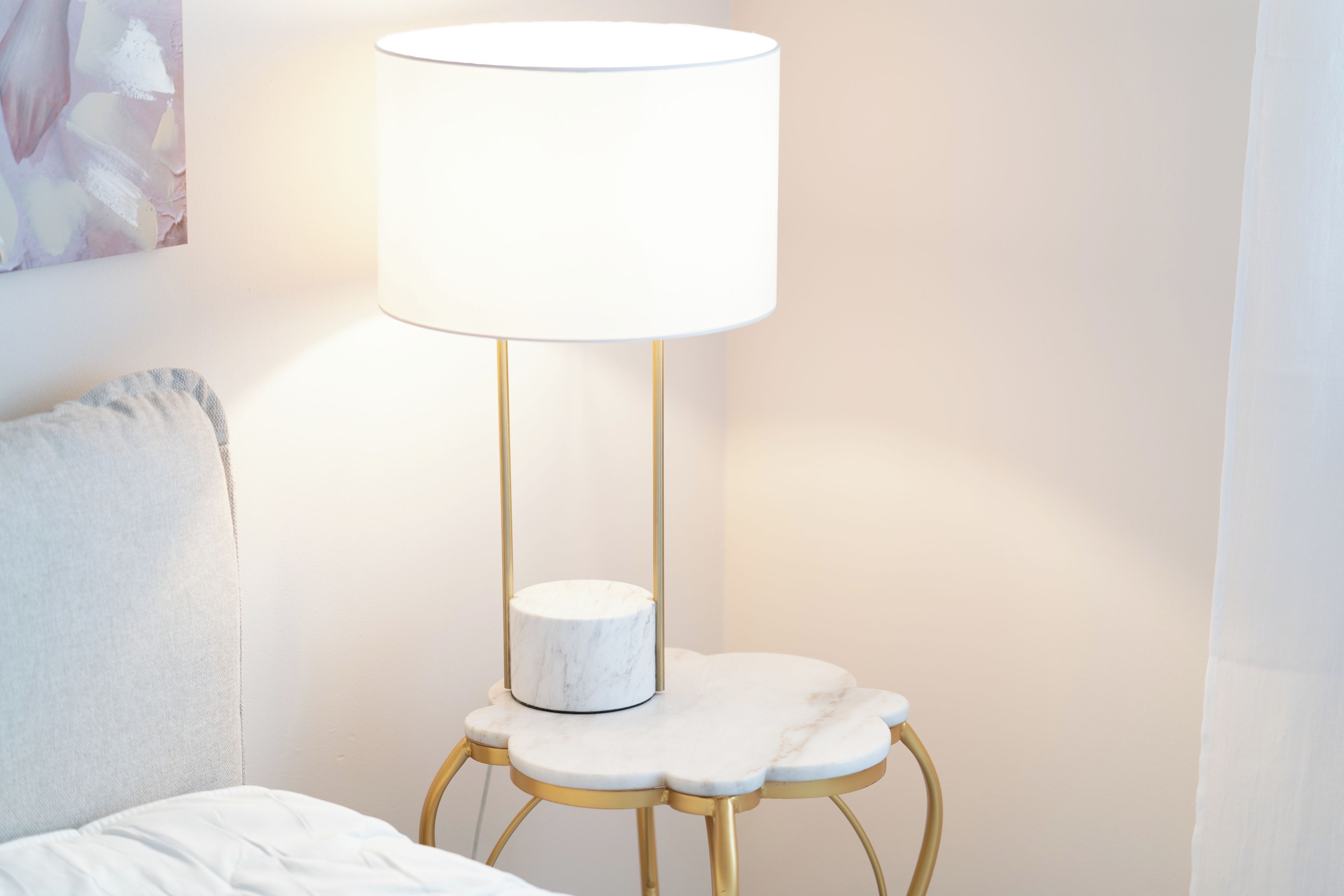 Een bed met een witte hoofdbord en een kussen, een klein rond tafeltje met een marmeren blad en een witte lampenkap, en een goudkleurige vloerlamp.