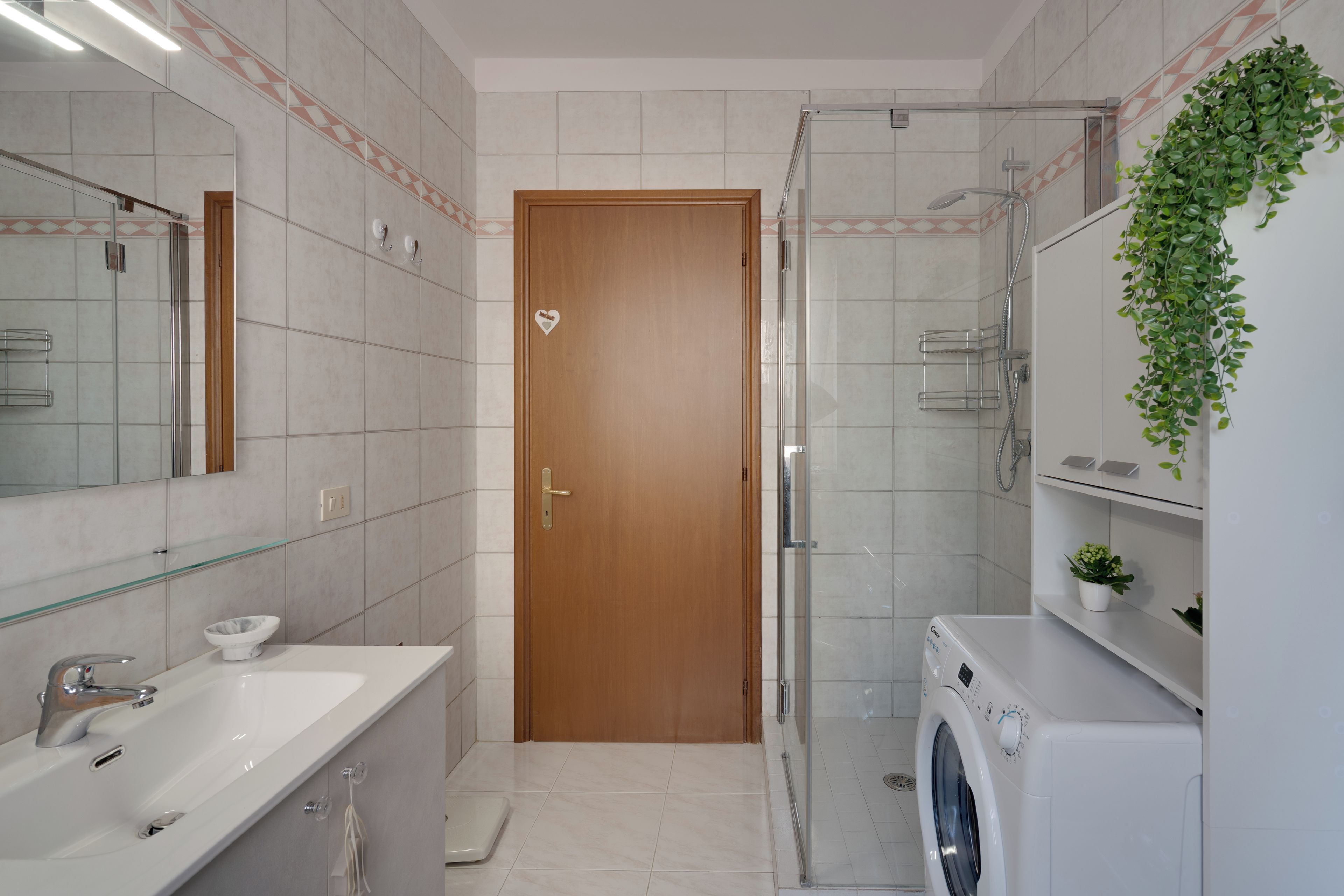Un bagno moderno con una porta in legno, un box doccia in vetro, una lavatrice bianca e uno specchio con ripiano in vetro sopra il lavandino.
