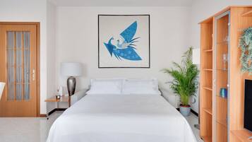 Une chambre d’hôtel avec un lit, une table de chevet, une lampe, une plante en pot et un meuble en bois doté d’étagères.