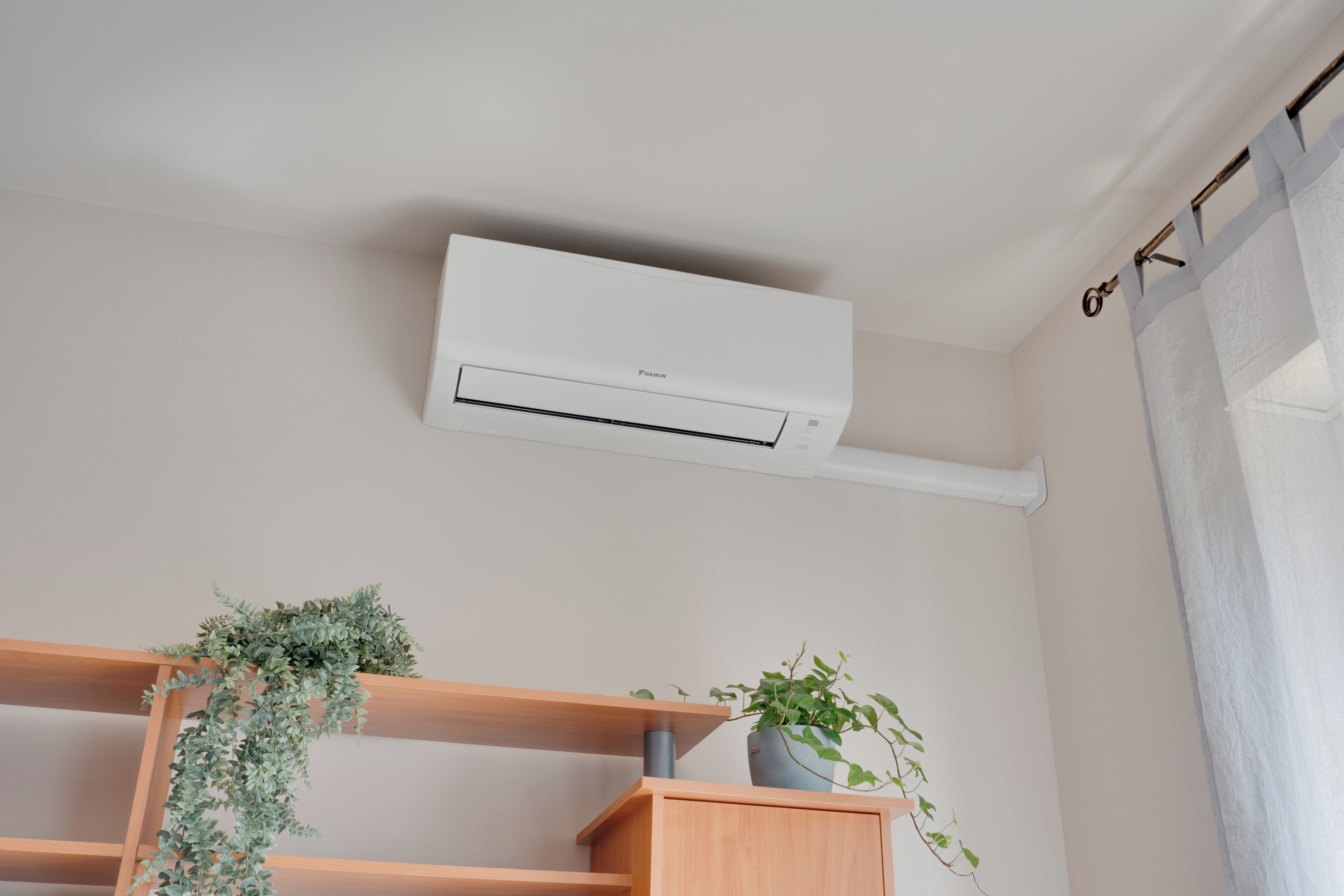 A room with a white air conditioning unit mounted on the wall, wooden shelves with plants, and a curtain on the window.