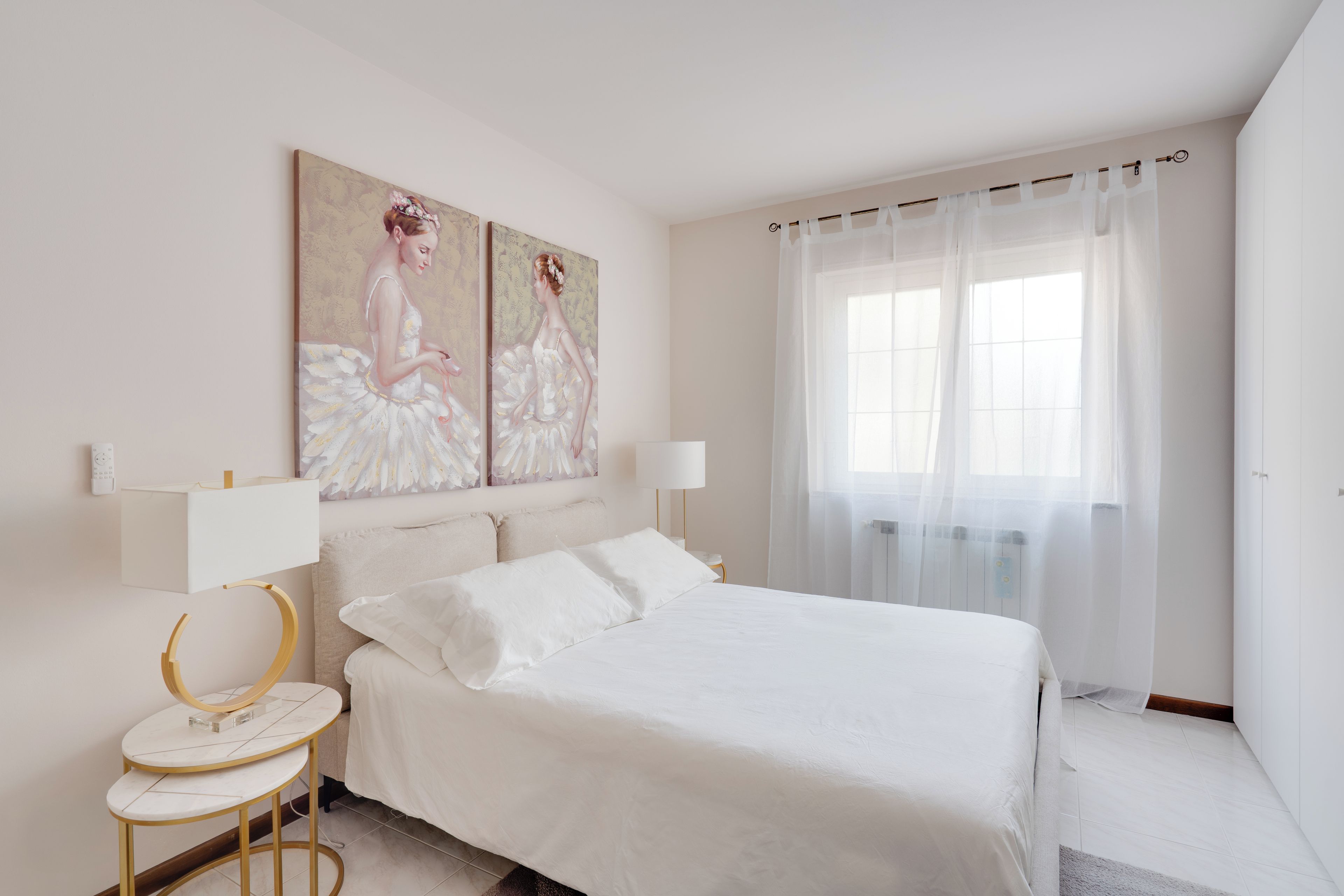 Un dormitorio con una cama, dos mesitas de noche, una lámpara de pared, un cuadro y un ventanal con cortinas translúcidas.