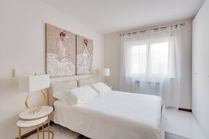 Un dormitorio con una cama, dos mesitas de noche, una lámpara de pared, un cuadro y un ventanal con cortinas translúcidas.
