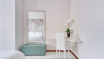 Un dormitorio con cama, un banco tapizado de color verde, un escritorio blanco con silla, una planta en maceta y un espejo grande que refleja una cama.