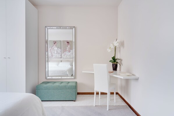Une chambre à coucher avec un lit, un banc recouvert d’un tissu vert, un bureau blanc avec une chaise, une plante en pot et un grand miroir qui reflète le lit.