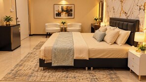 Un dormitorio moderno con una cama grande, mesitas de noche, un pequeño escritorio, dos sillas, una planta en maceta y una obra de arte colgada en la pared.