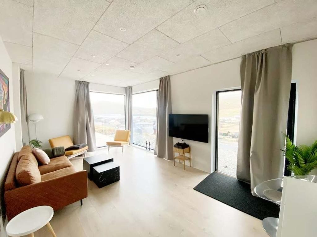 Una sala moderna con sofá, mesa de centro, televisor de pantalla plana y un ventanal con cortinas.