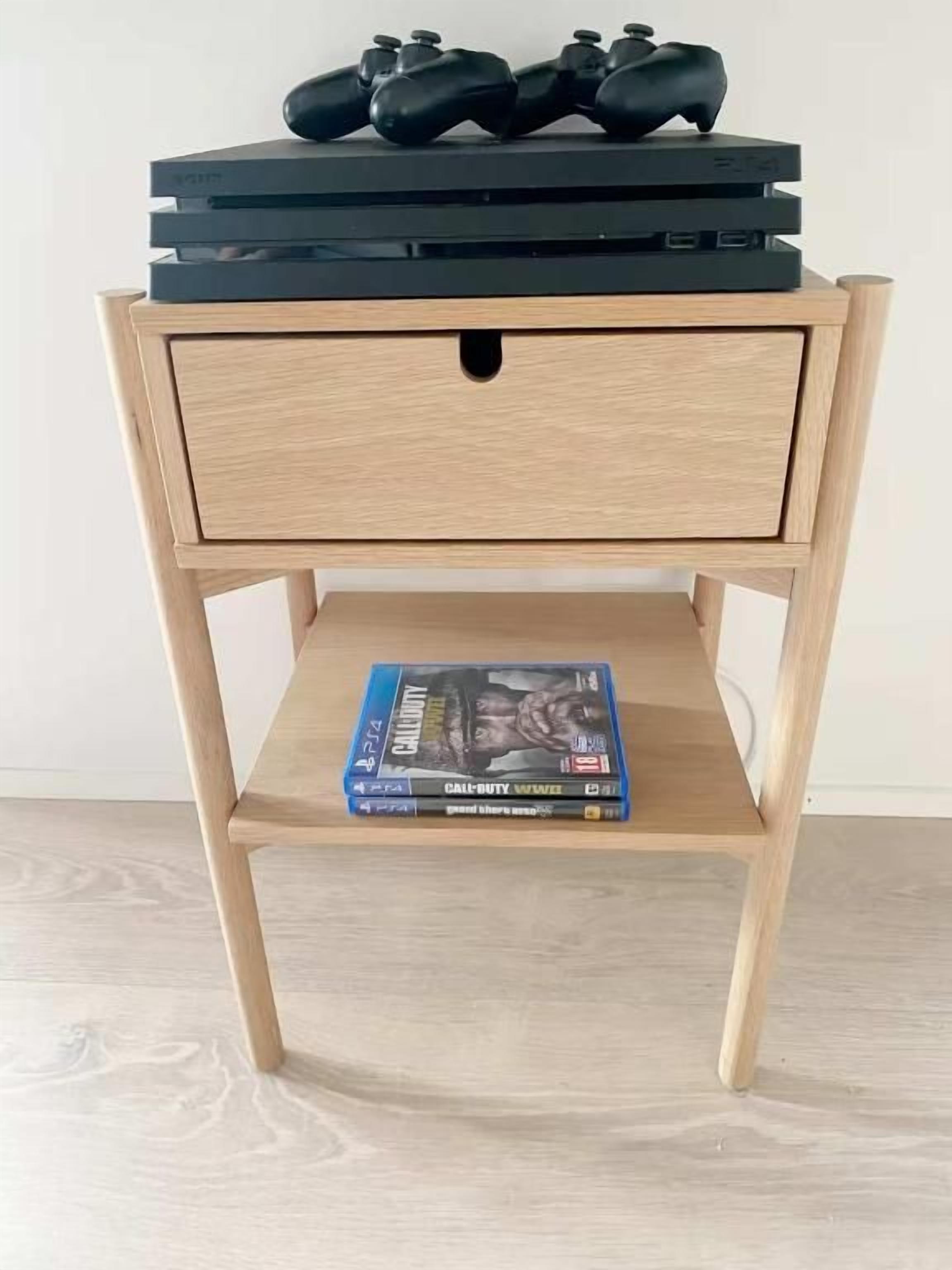 Una mesita de noche de madera con un cajón y un estante, que sostiene una consola de videojuegos y su estuche.