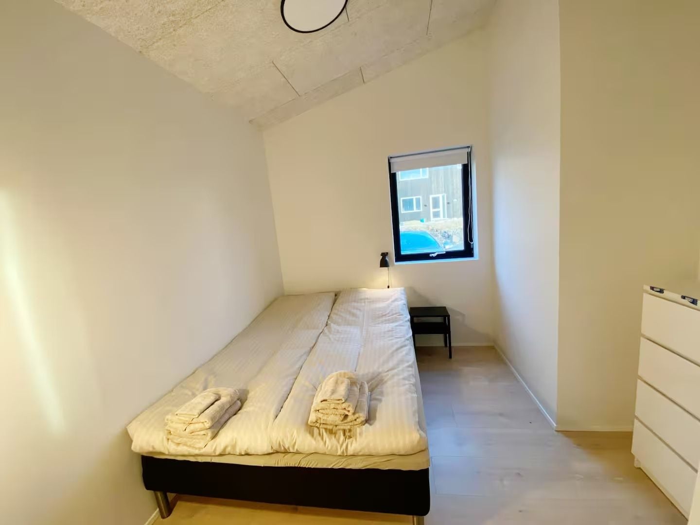 A small, single-bed room with a window, a bedside table, and a dresser.