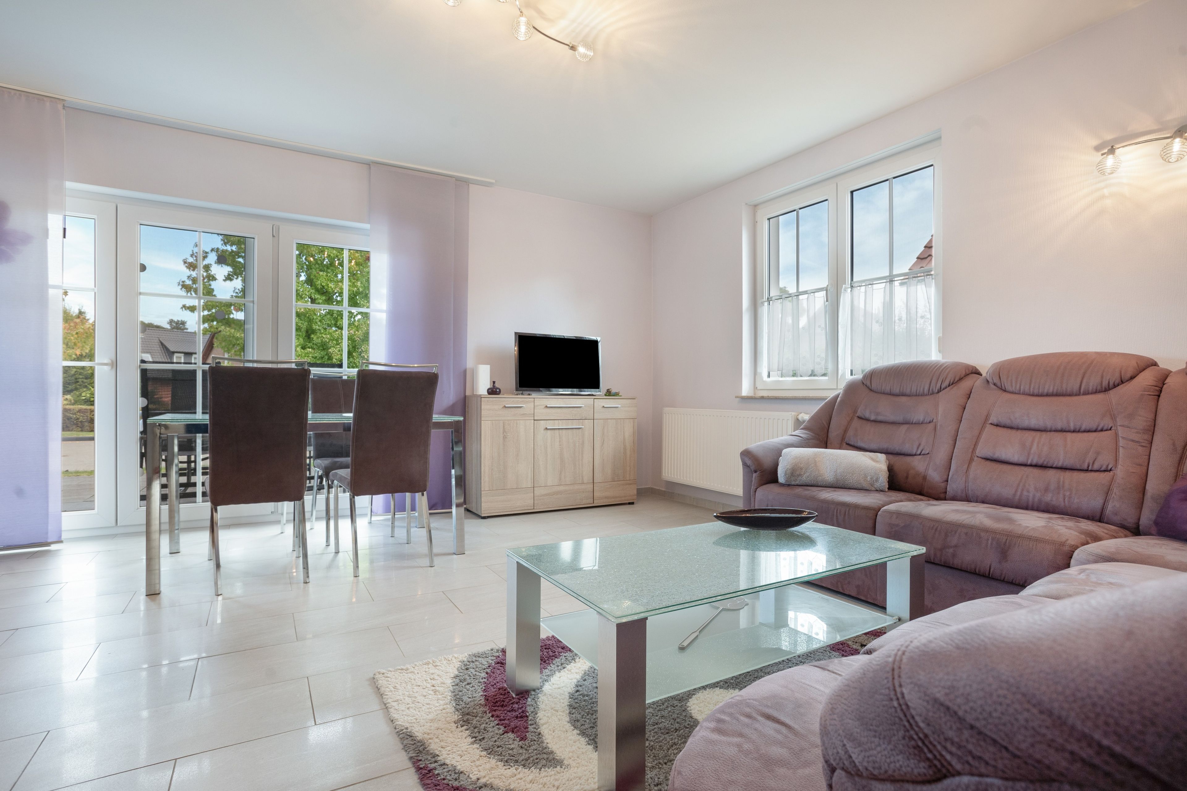 Een woonkamer met een glazen salontafel, een bank, een televisie en een eethoek met stoelen.