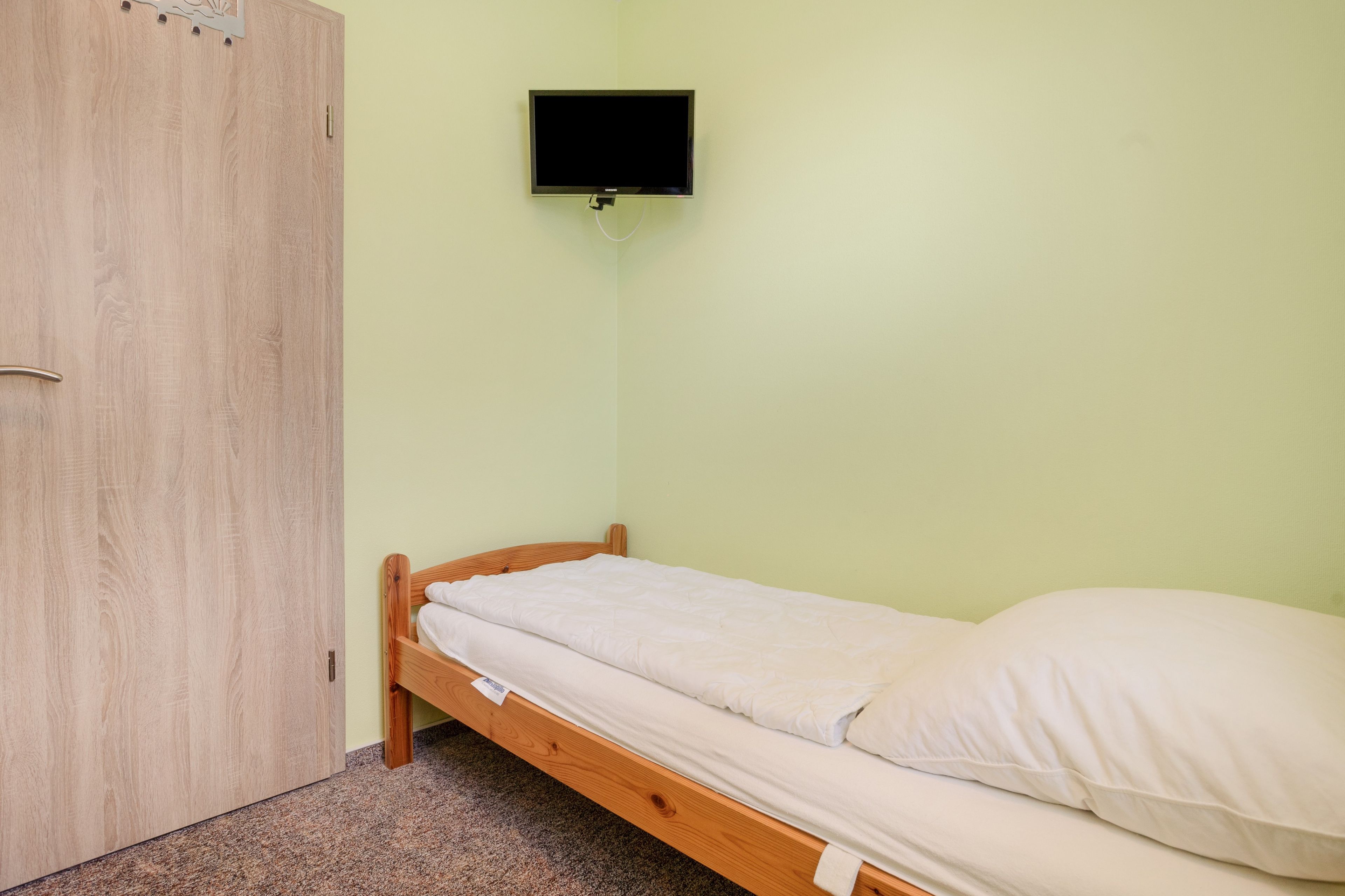 Een kleine kamer met een enkel bed, een houten bedframe en een aan de muur bevestigde televisie.
