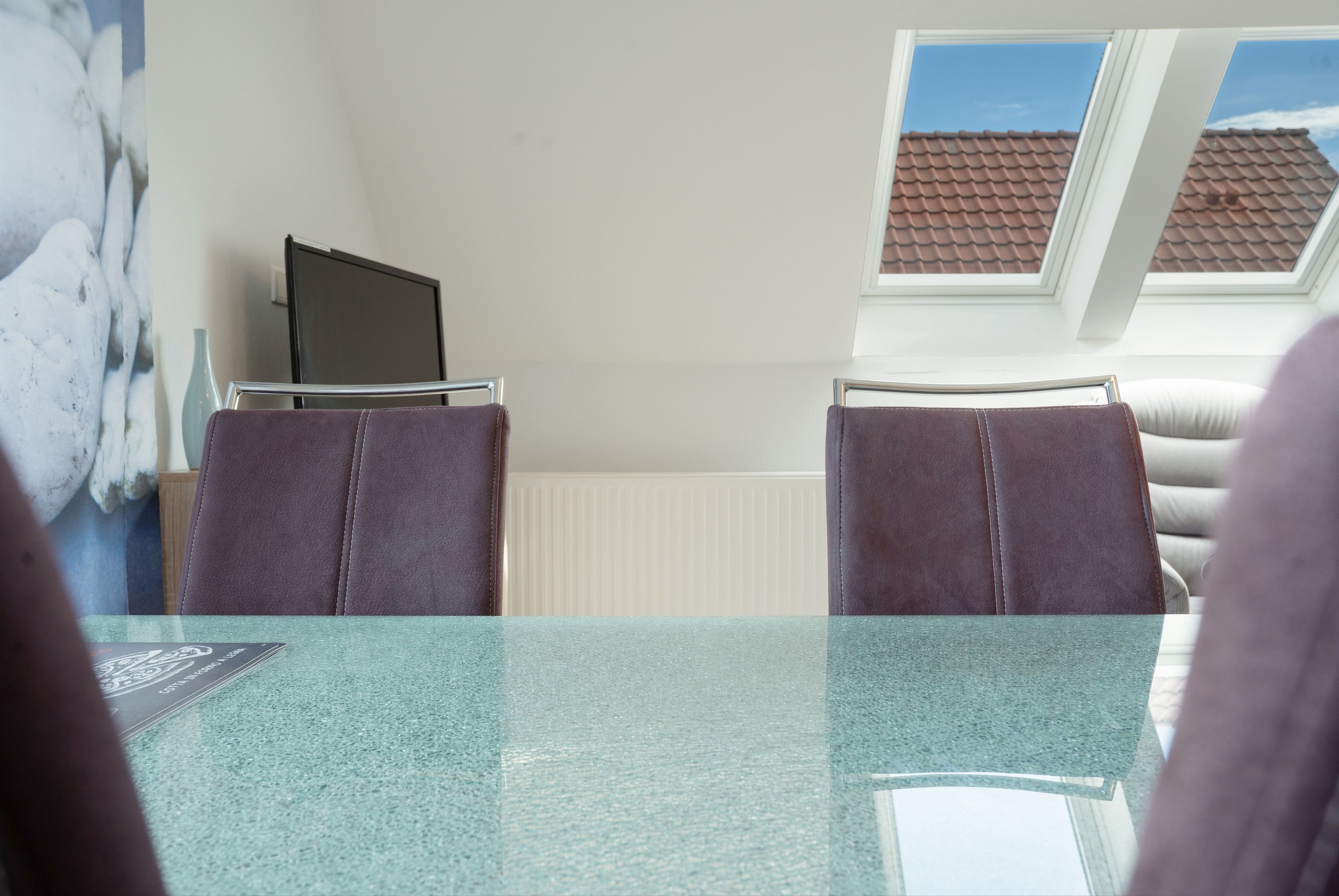 Ein Zimmer mit einem Glastisch, zwei Stühlen, einem Laptop auf einem Ständer, einem Heizkörper und einem Fenster mit Blick auf ein Ziegeldach.