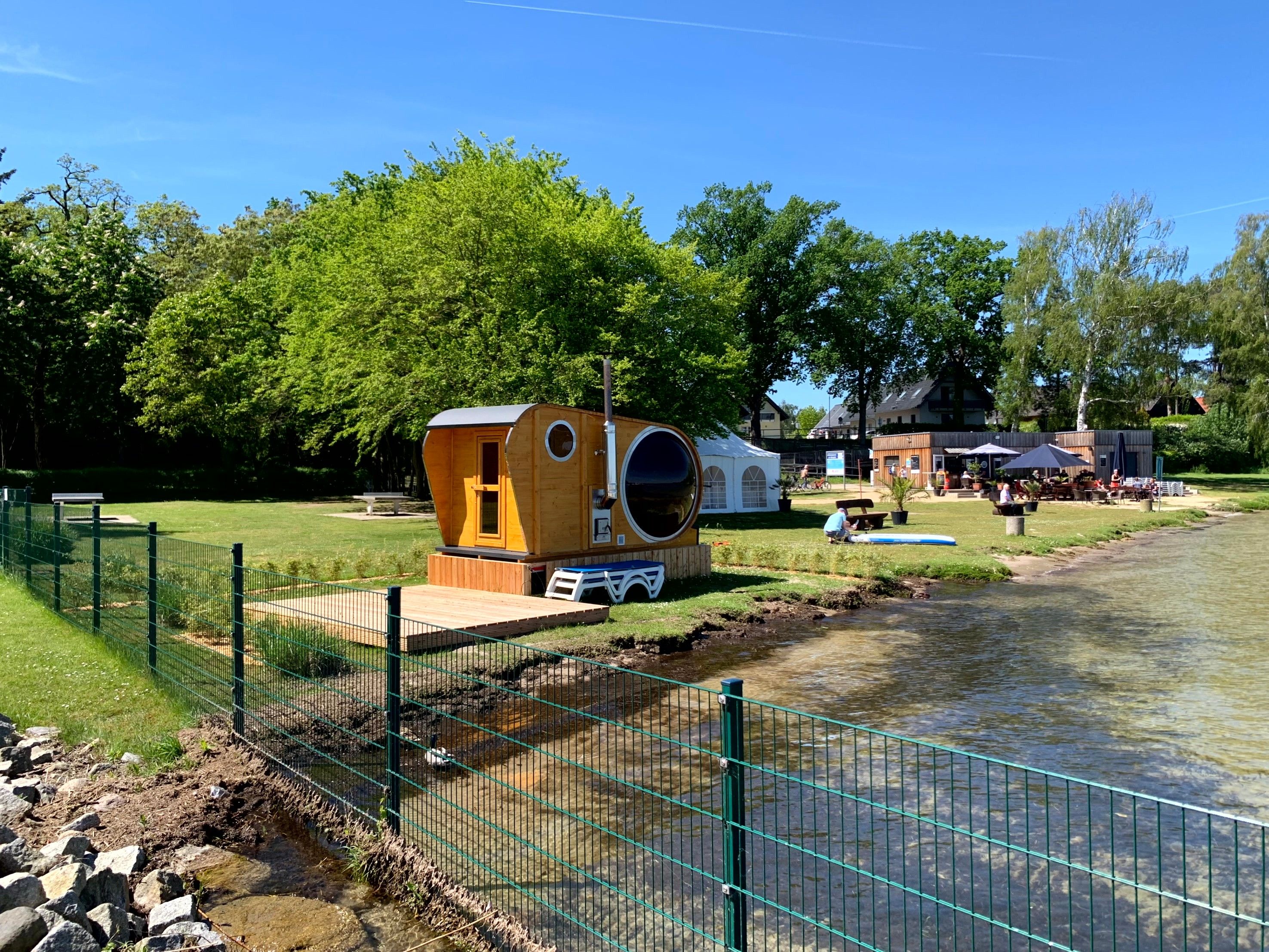 A wooden cabin with a large circular window, situated by a river with a grassy area and a fence in the foreground.