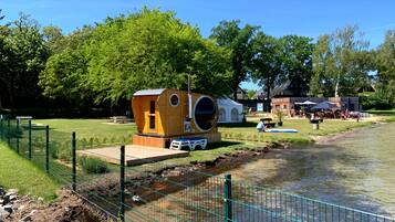 A wooden cabin with a large circular window, situated by a river with a grassy area and a fence in the foreground.