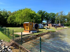A wooden cabin with a large circular window, situated by a river with a grassy area and a fence in the foreground. - Apartment 'U 6 OG Biber' with Sea View, Shared Garden and Wi-Fi (Röbel/Müritz)