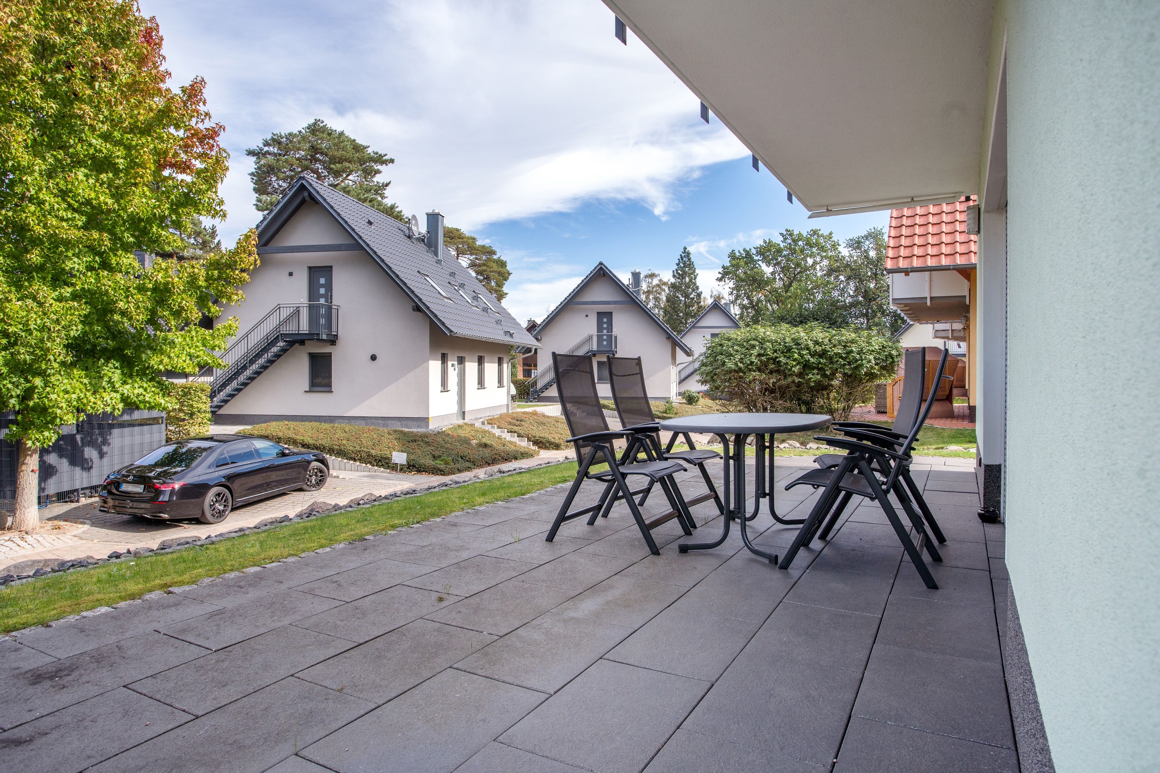 Eine Terrassenfläche mit Tisch und Stühlen, ein geparktes Auto in der Nähe und Häuser im Hintergrund.