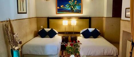 A hotel room with two beds, a nightstand, a vase with flowers, and a painting on the wall.