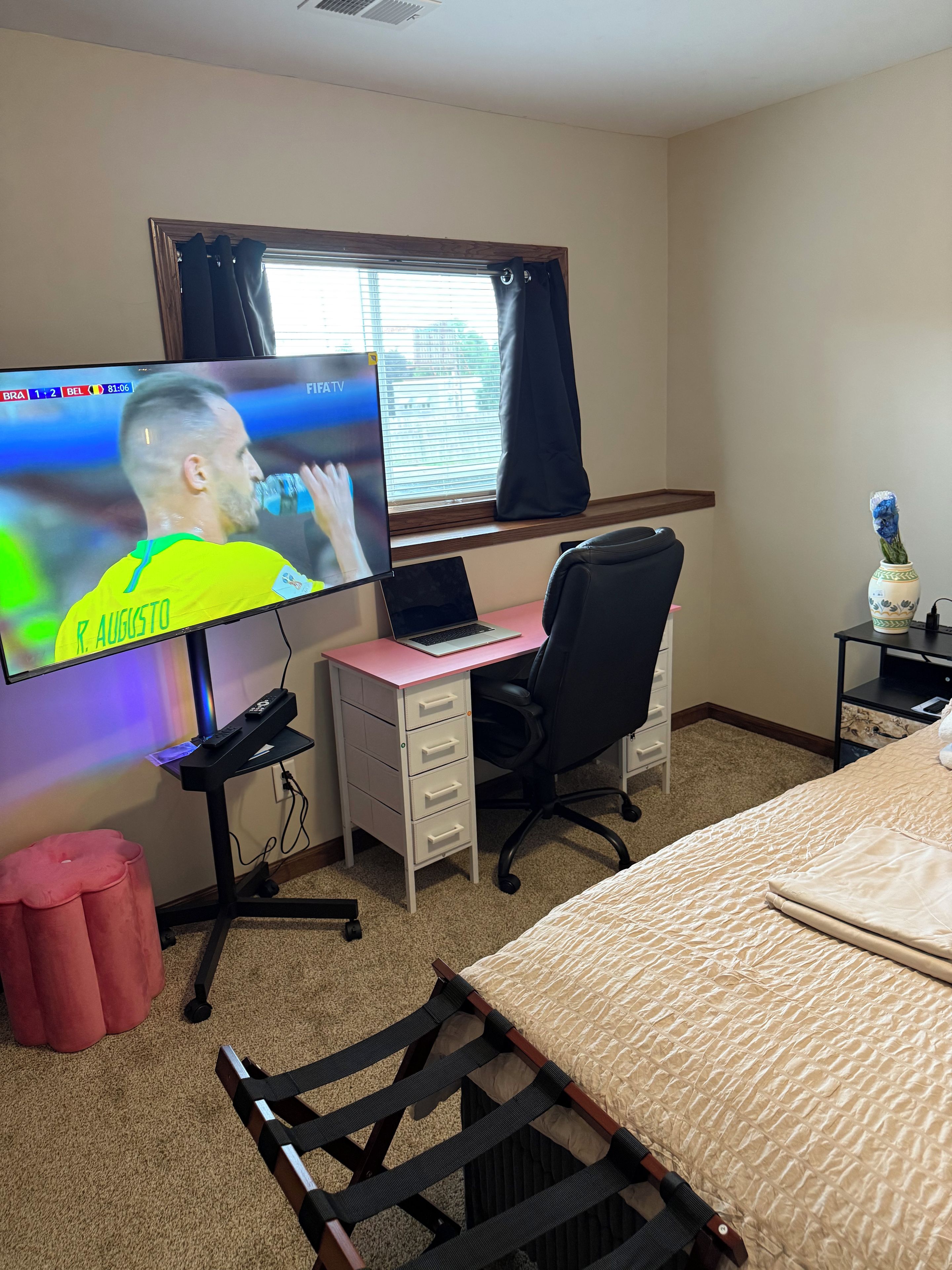 A hotel room with a bed, a desk with a laptop, a TV displaying a sports event, a chair, a small table with a vase, and a window with curtains.