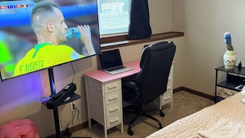 A hotel room with a bed, a desk with a laptop, a TV displaying a sports event, a chair, a small table with a vase, and a window with curtains.