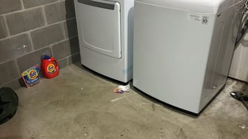 Two white front-loading washing machines in a laundry area with a concrete floor, a brick wall, and a door in the background.