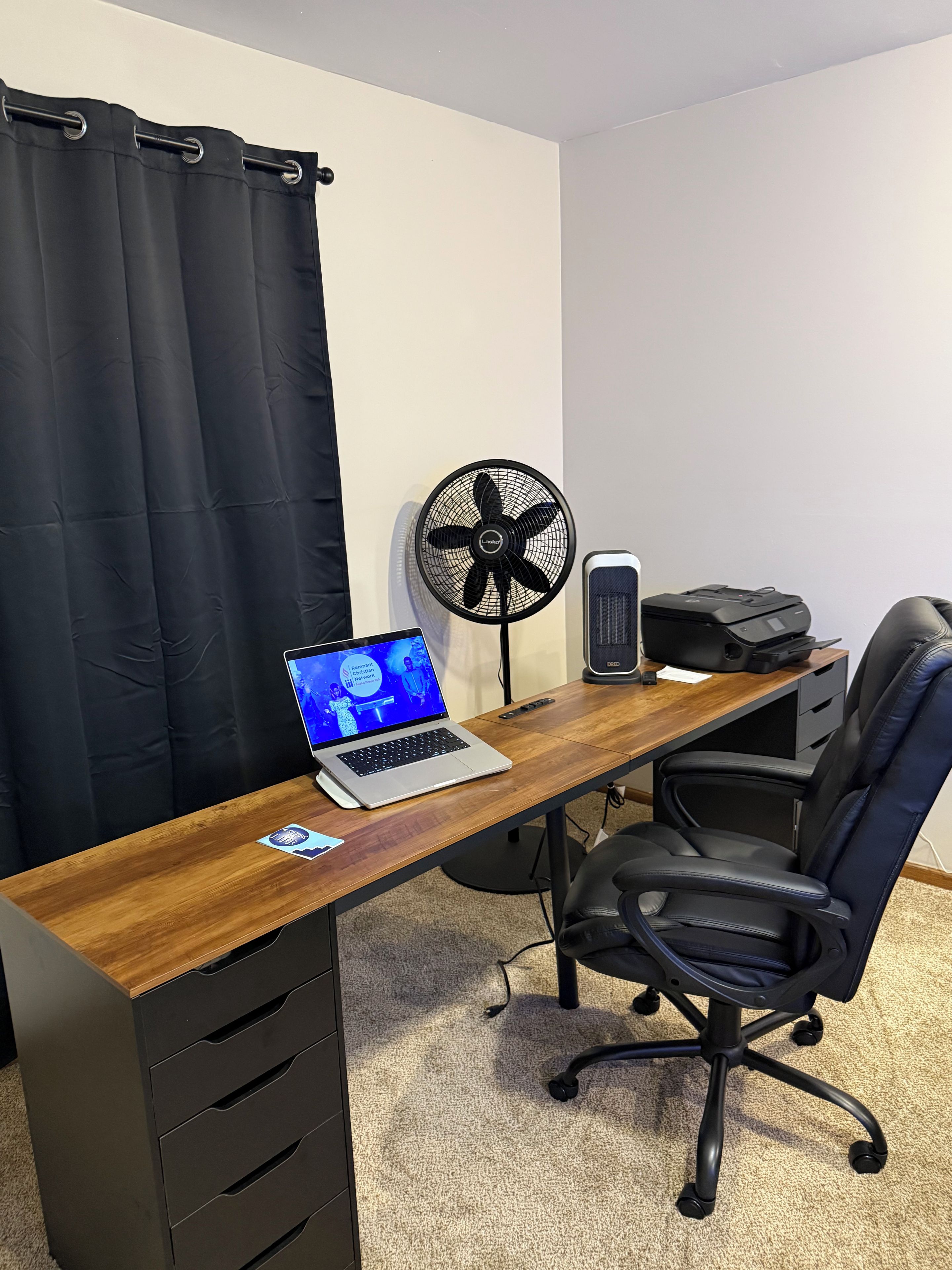 Un espace de travail comprenant un bureau en bois, un ordinateur portable, une chaise de bureau noire, un ventilateur sur pied et une imprimante.