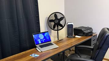 Un espace de travail comprenant un bureau en bois, un ordinateur portable, une chaise de bureau noire, un ventilateur sur pied et une imprimante.
