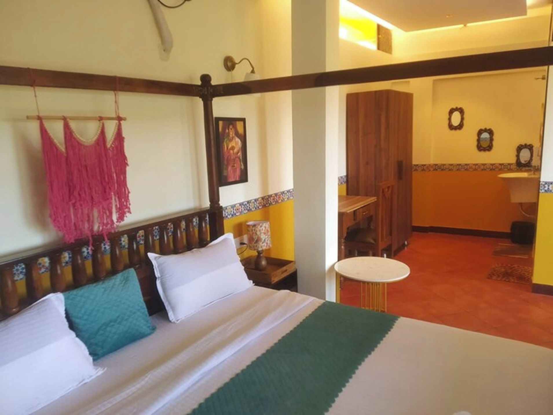 A hotel room with a bed, a hanging macrame piece, a painting, and a bathroom visible through an open door.