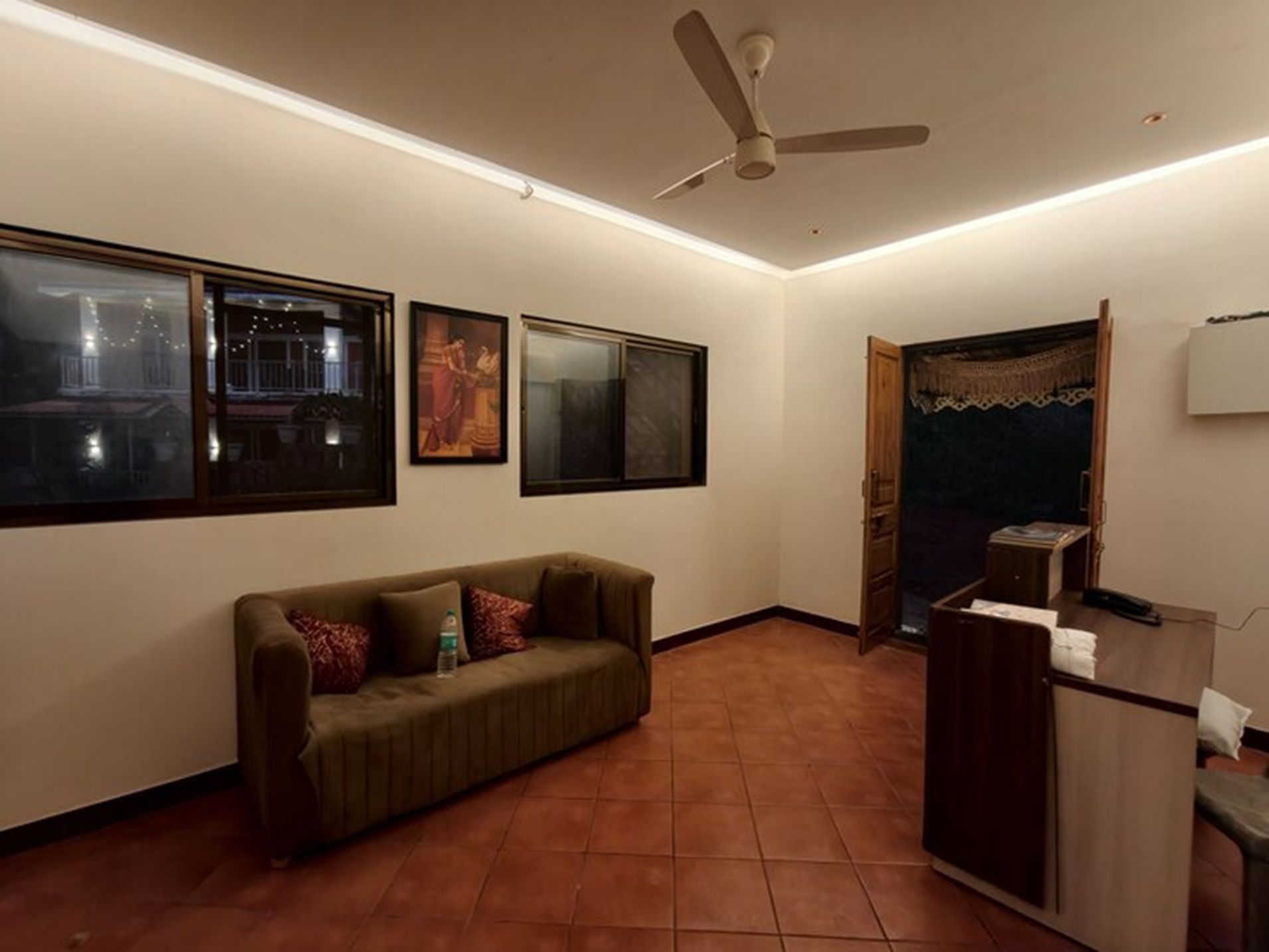 Una habitación con ventilador de techo, sofá, escritorio y ventana con vista a un edificio de noche.