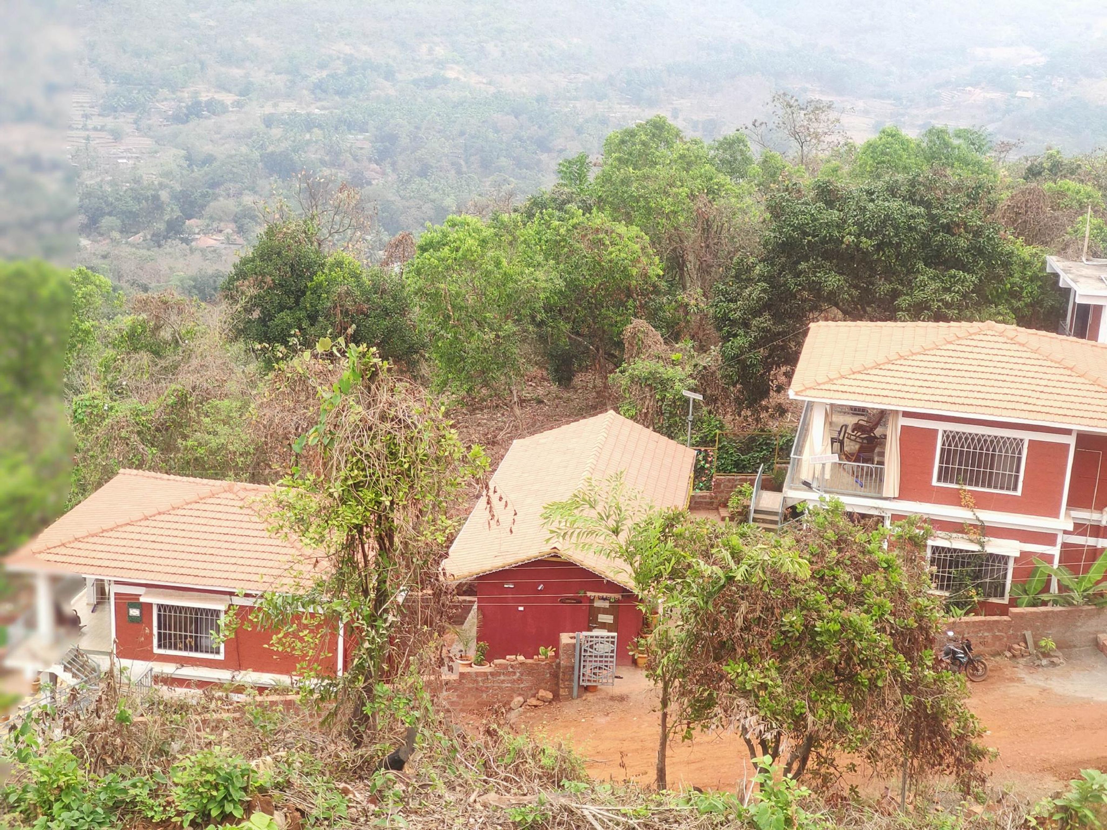 A residential area with houses featuring red walls and tiled roofs, surrounded by greenery and hilly terrain.