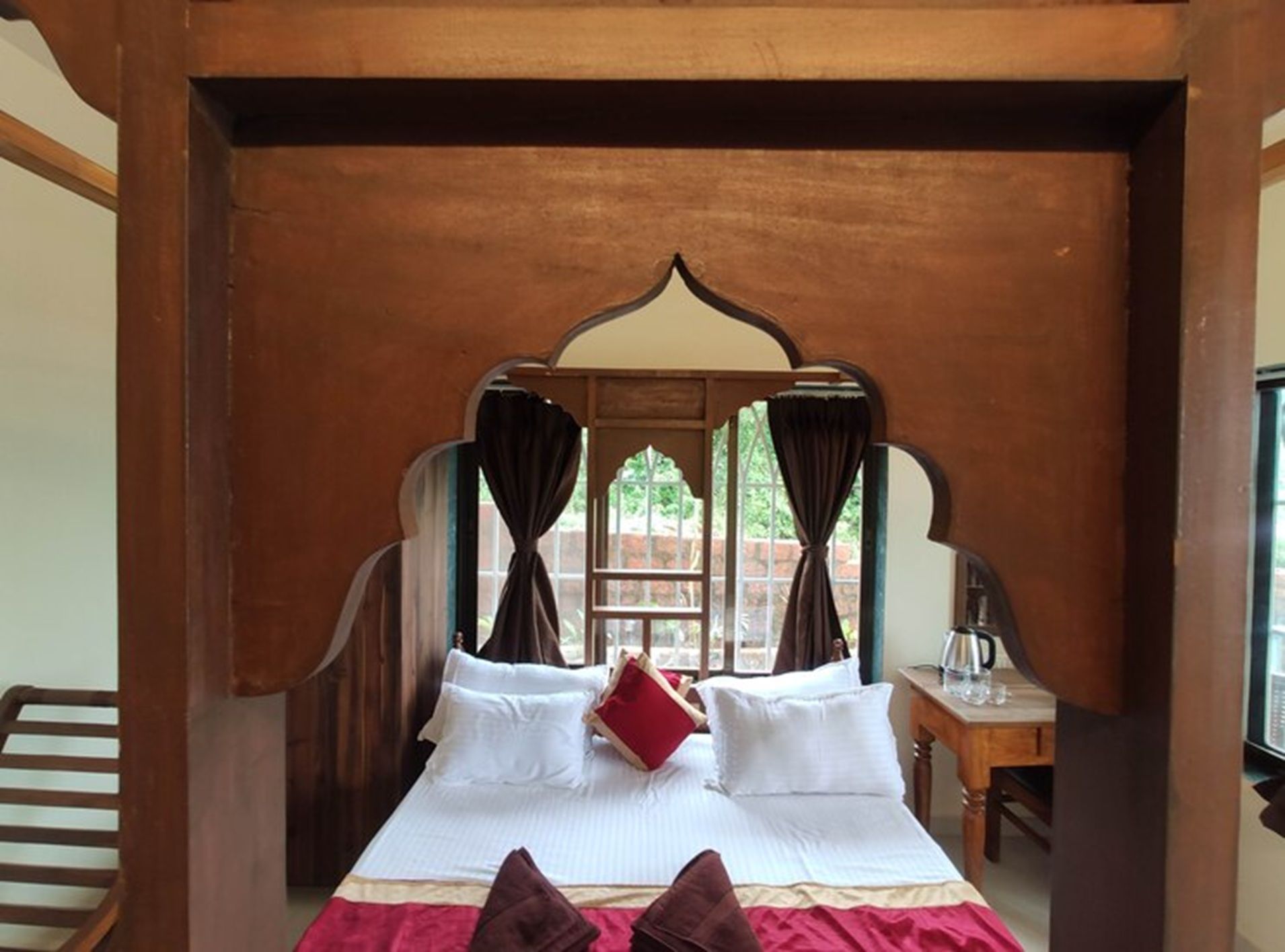 A bed with white linens and a red pillow, a wooden headboard, and a window with curtains.