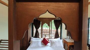 A bed with white linens and a red pillow, a wooden headboard, and a window with curtains.