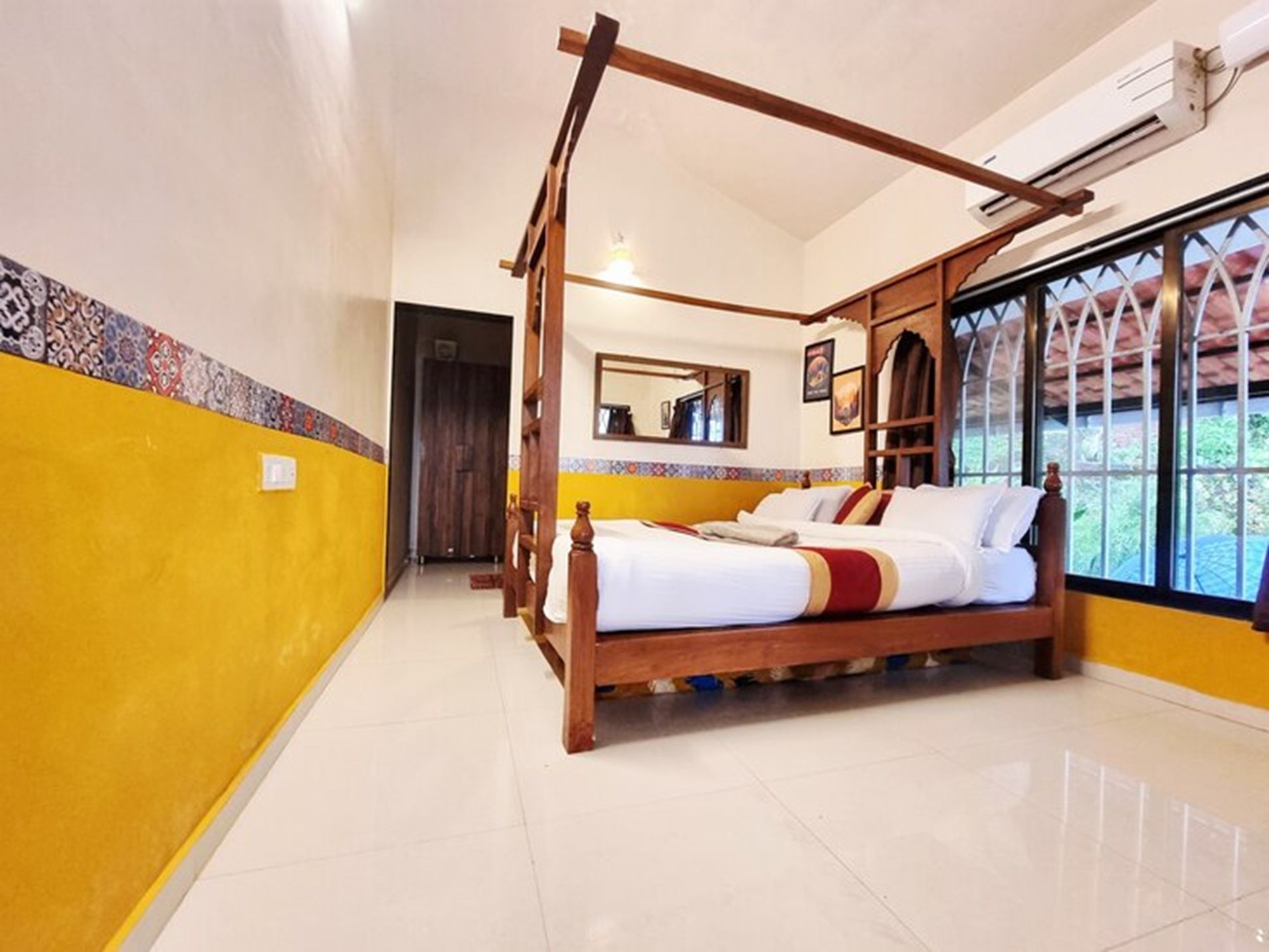 A bedroom with a four-poster bed, a large window with decorative ironwork, and a tiled floor.