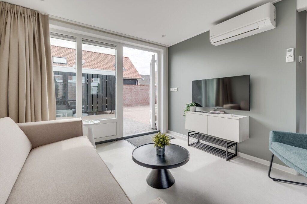 A modern living room with a sofa, a round coffee table with a plant, a TV mounted on the wall, and a sliding glass door leading to an outdoor area.