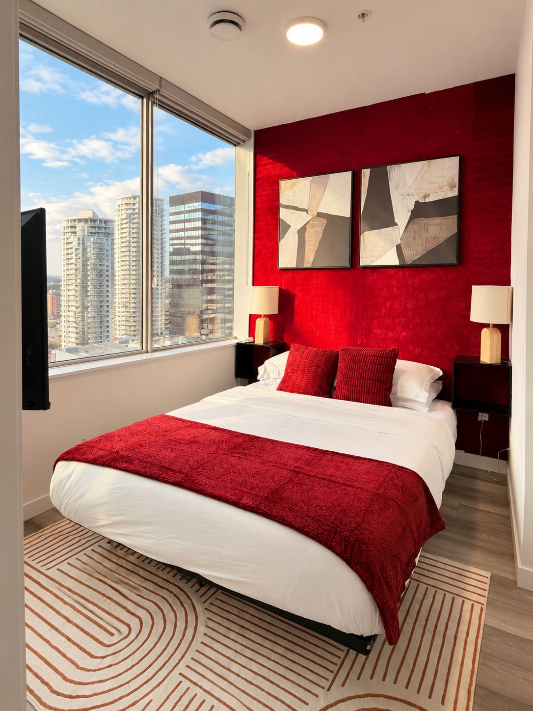 A modern bedroom with a large bed, red accent walls, two abstract paintings, a city view, and a cityscape visible through the window.
