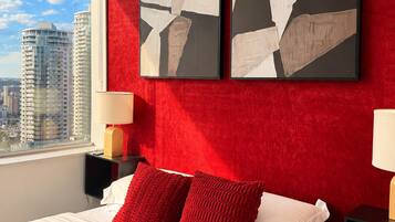 A modern hotel room with a red accent wall, a large bed with white and red bedding, two side tables with lamps, and a city view.