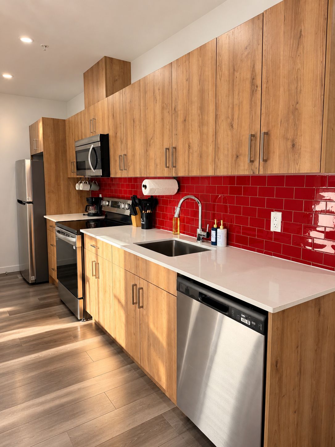 A modern kitchen with wooden cabinets, stainless steel appliances, a red tiled backsplash, and a white countertop.